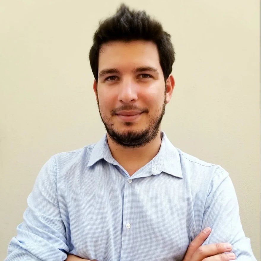 Evangelos Mitsokapas, arms folded, in a light blue shirt against a warm neutral wall.